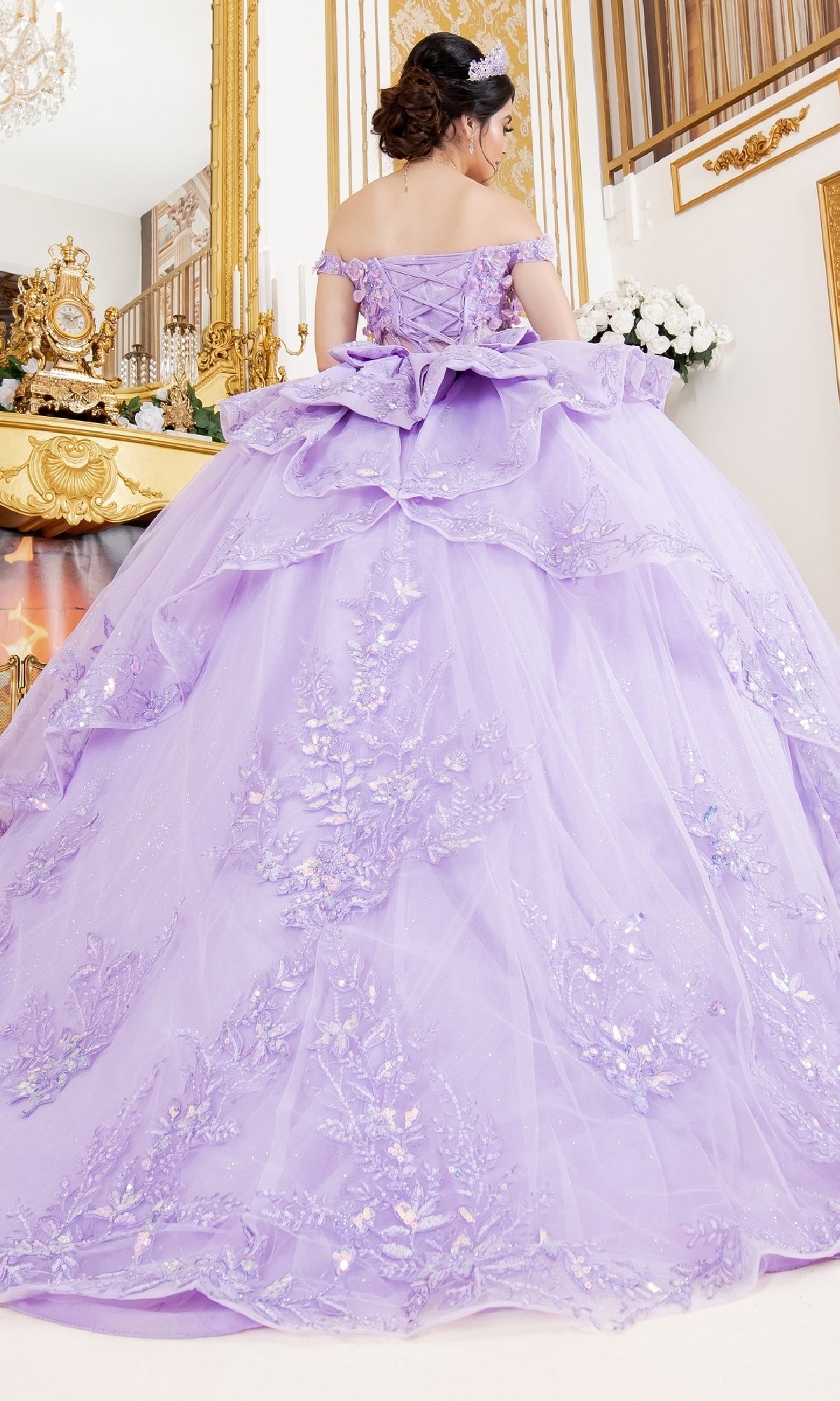 A woman stands indoors in an ornate room with gold accents and a large mirror, wearing the Cinderella Couture 8160J—an off-the-shoulder lavender quinceanera ball gown with embroidered lace appliques.